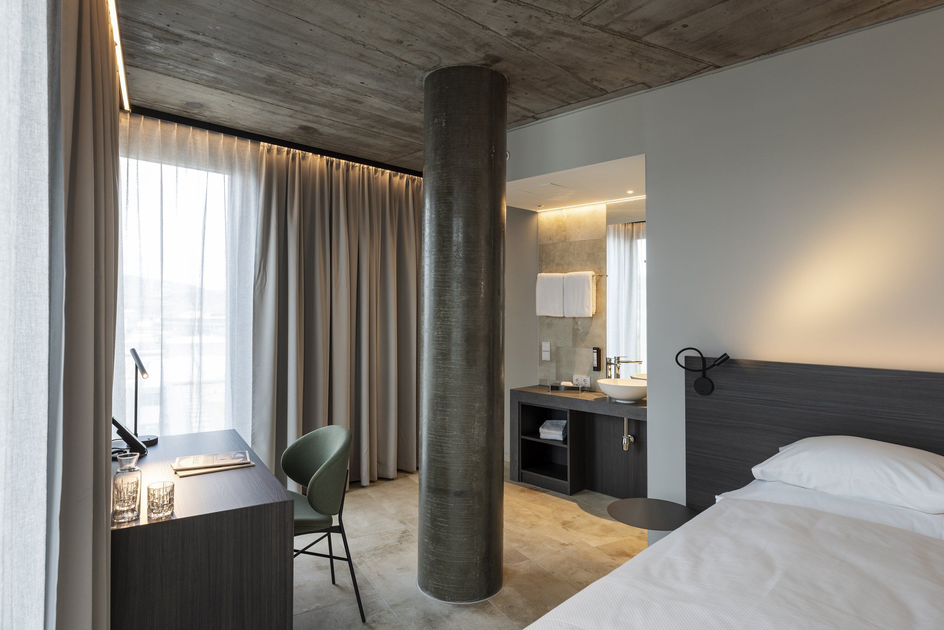 Una habitación de hotel con escritorio, silla, cama y un gran pilar cilíndrico.