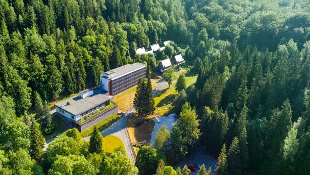 Une vue aérienne d’un bâtiment entouré d’une forêt dense, avec un espace de stationnement et quelques petites constructions à proximité.