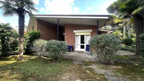 Un edificio con fachada de ladrillo, un acceso cubierto y un pequeño jardín con diversas plantas y una palmera.