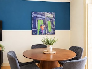 A dining area with a round wooden table, four upholstered chairs, a potted plant, and a colorful abstract painting on the wall. - Sao Bento Tramview Residence (Lisboa)
