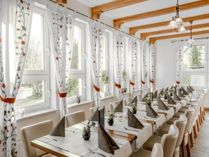 Ein langer Esstisch mit mehreren Stühlen, weiße Vorhänge mit Blumenmuster und Holzbalken an der Decke.