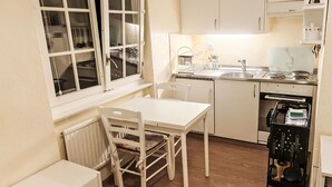 Cocina compacta con muebles blancos, una mesa de comedor y sillas. Se ve una ventana con cortinas, y un electrodoméstico en la encimera.