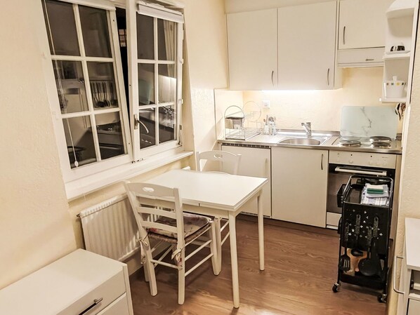 Eine kleine Küche mit weißen Schränken, einem Esstisch und Stühlen. Ein Fenster mit Vorhängen ist sichtbar, und ein kleines Gerät steht auf der Arbeitsplatte.