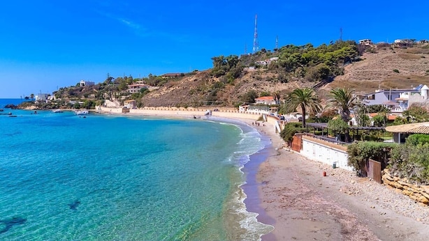 Une plage côtière aux eaux turquoise et limpides, avec un rivage sablonneux et des bâtiments construits sur une colline.