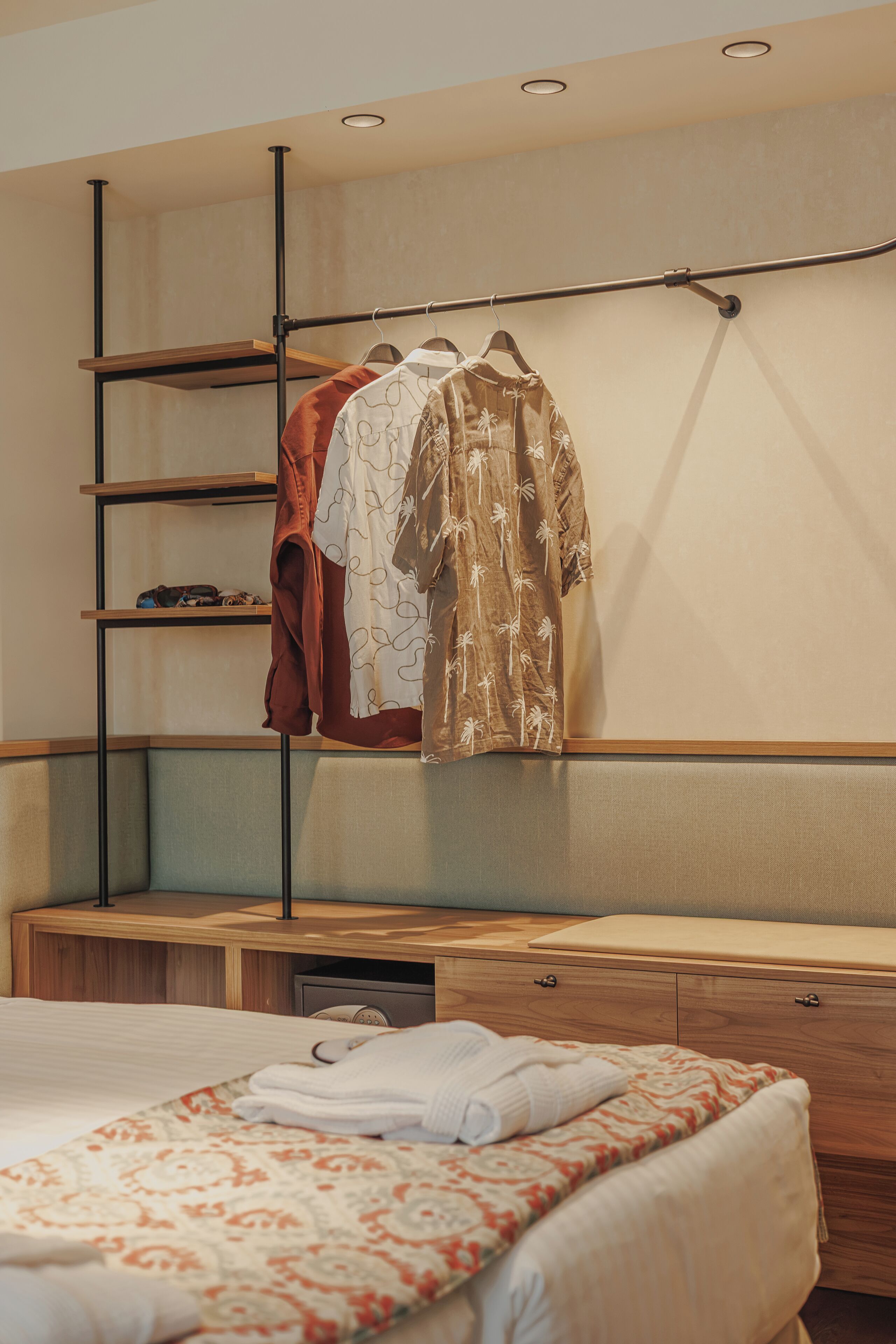 Een hotelkamer met een bed, een houten nachtkastje, een inbouwkast en een kledingroede met kleding.