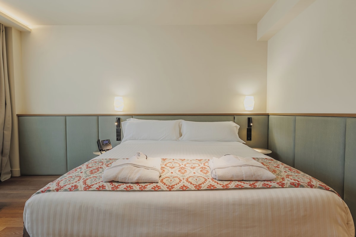 Una habitación de hotel con cama, dos lámparas de noche, un teléfono y una ventana con cortinas.