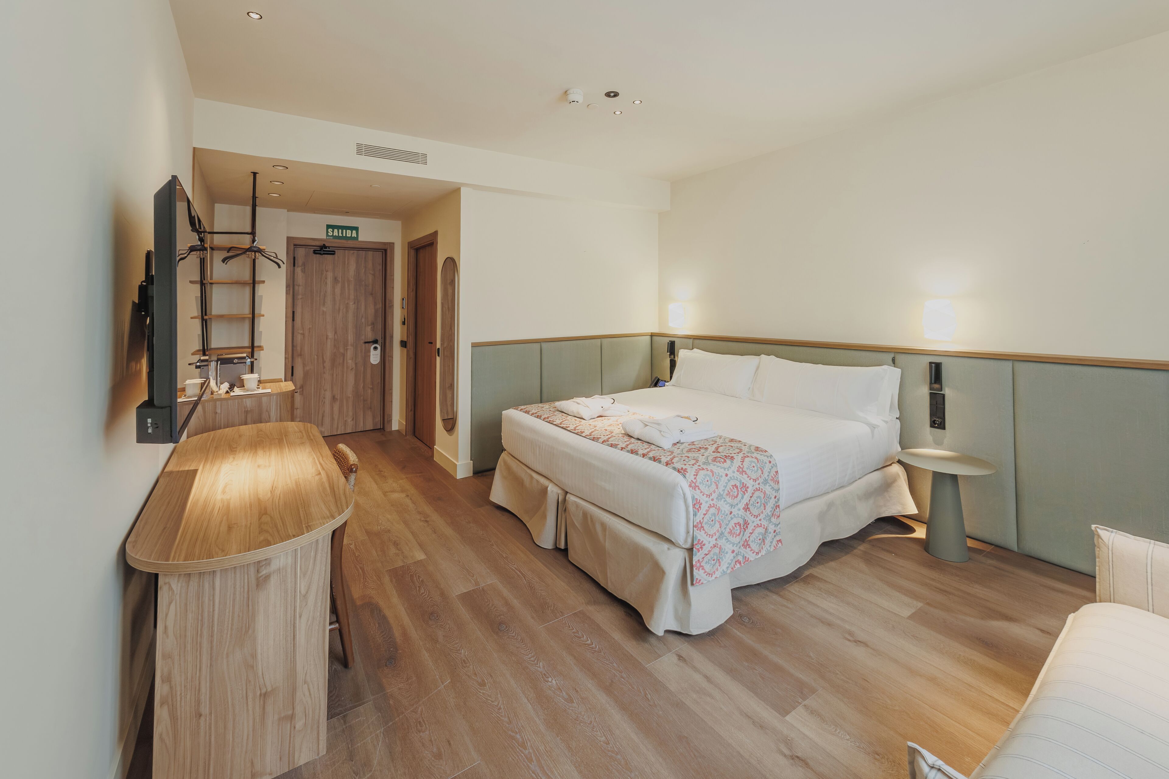A hotel room with a bed, bedside tables, a desk, a chair, a wardrobe, and a mirror.