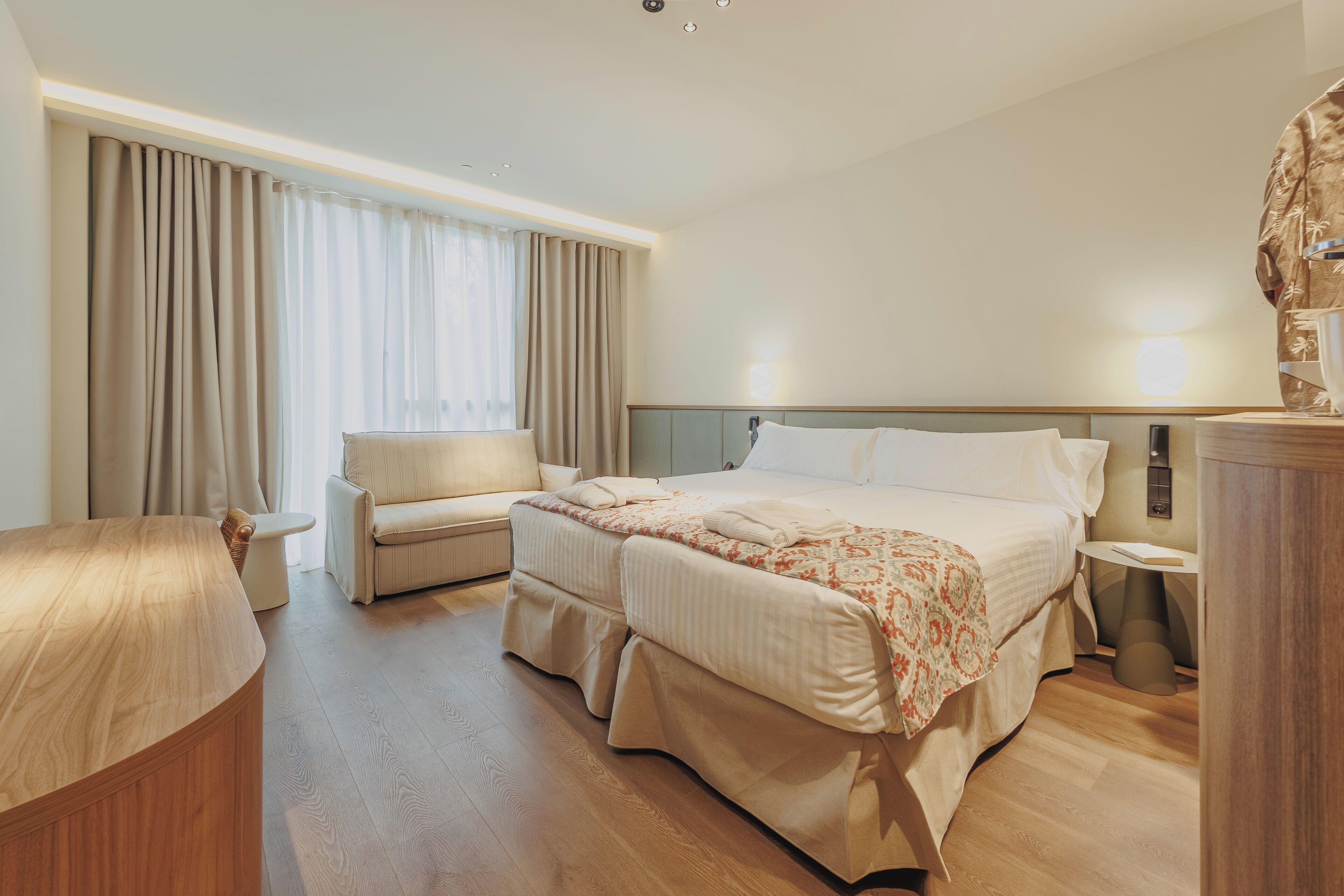 Una habitación de hotel con cama, una silla, una mesita de noche y un ventanal con cortinas.
