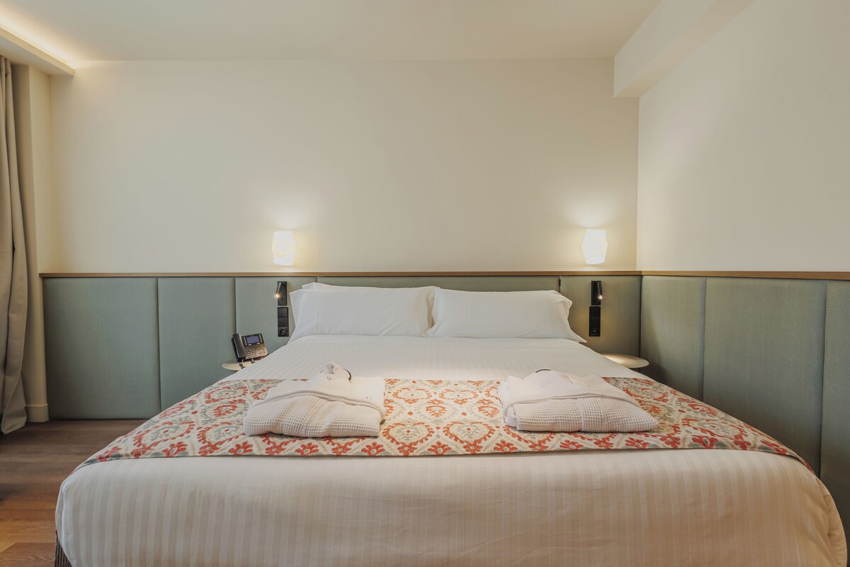 A hotel room with a bed, two bedside lamps, a telephone, and a window with curtains.