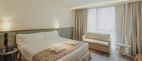 Habitación de hotel con cama, mesita de noche, un pequeño sofá, ventana con cortinas y teléfono junto a la cama.