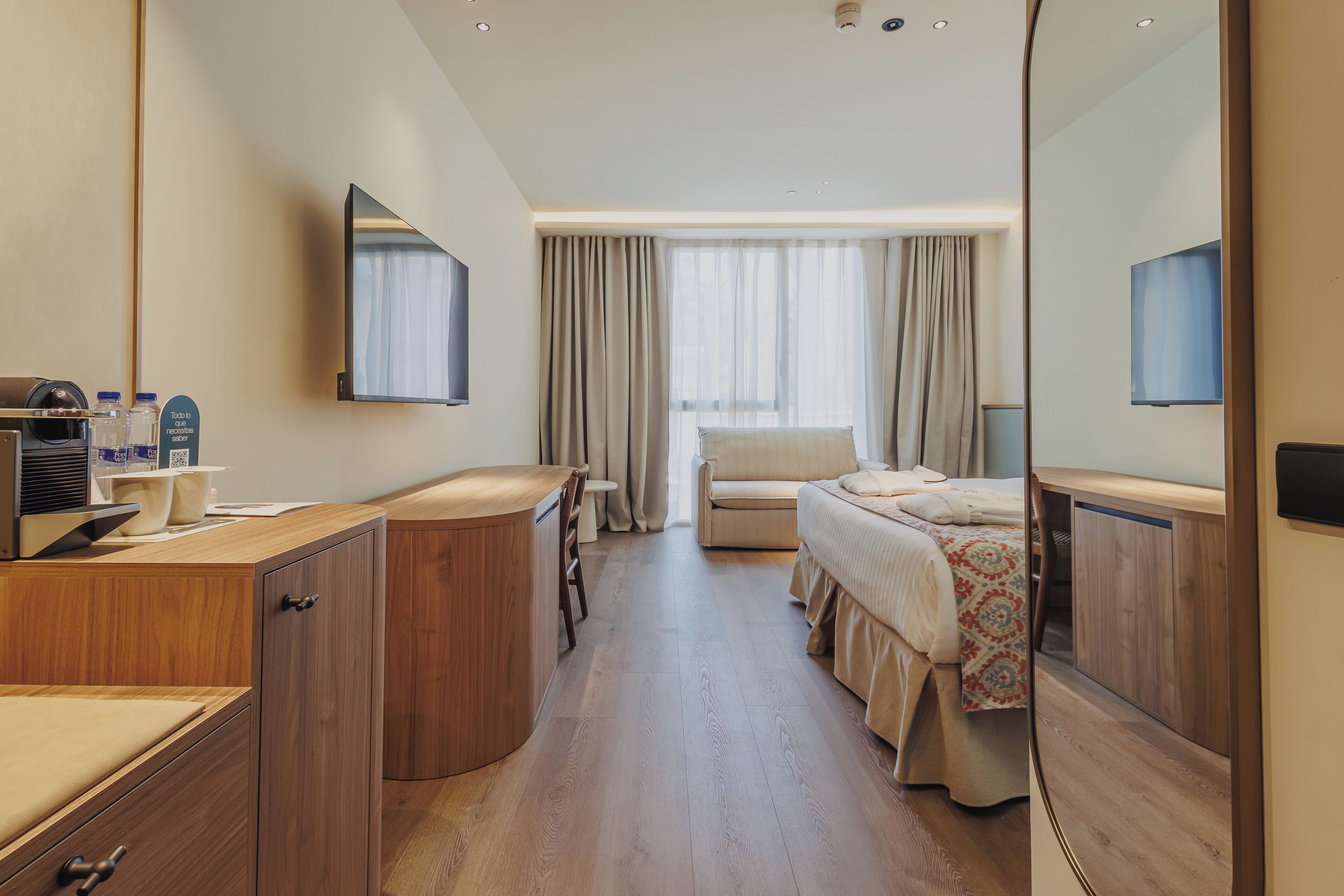 Habitación de hotel con escritorio de madera, cama con colcha estampada, sofá, televisor y un espejo grande.