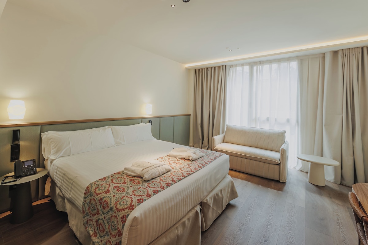 Habitación de hotel con cama, mesita de noche, un pequeño sofá, ventana con cortinas y teléfono junto a la cama.