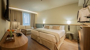Una habitación de hotel con una cama, un escritorio con una computadora portátil, una silla, un televisor montado en la pared y una pequeña mesa con una botella y una cámara.