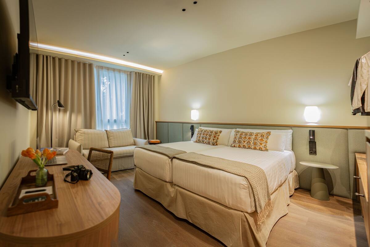 A hotel room with a bed, a desk with a laptop, a chair, a TV mounted on the wall, and a small table with a bottle and a camera.