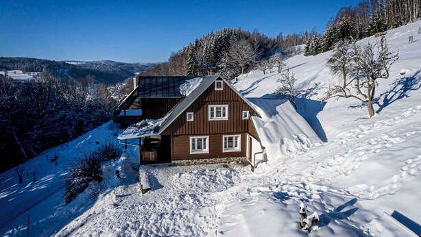 Una cabaña de madera con techo inclinado, rodeada de colinas y árboles cubiertos de nieve.