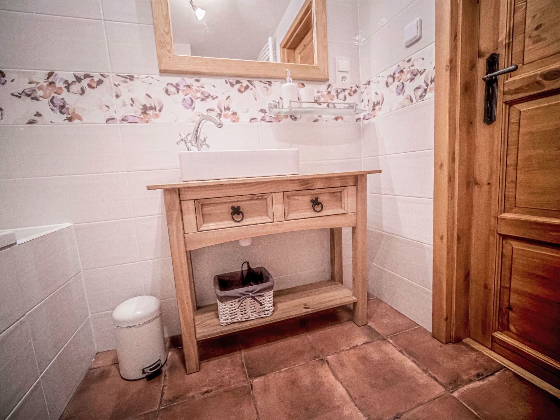 A bathroom with a wooden vanity, a white sink, a mirror, and a trash bin.