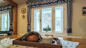 Una habitación acogedora con suelo de madera, un ventanal con cortinas a cuadros y una mesa con una cesta de pan y una tetera.