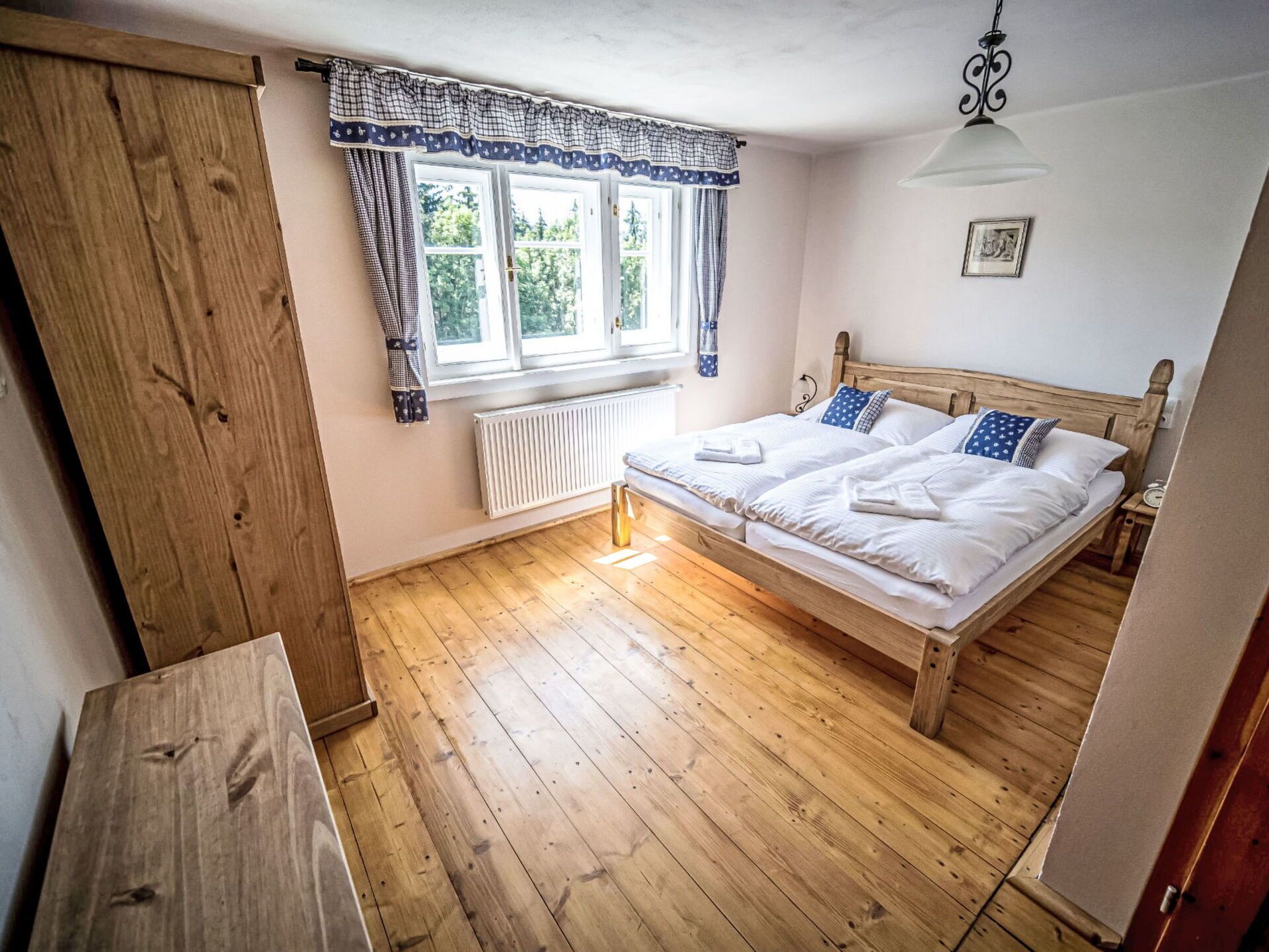 Un dormitorio con suelo de madera, una cama doble con ropa blanca, una ventana con cortinas, un radiador y un armario de madera.