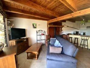 A living room with a television, sofa, wooden coffee table, and a kitchen area with a bar and stools. - House on the Edge of the Forest – Sleeps 8 (Chassignolles)