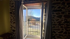 A room with a stone wall, a wooden floor, a window with curtains, and a view of a fenced outdoor area with trees and hills.