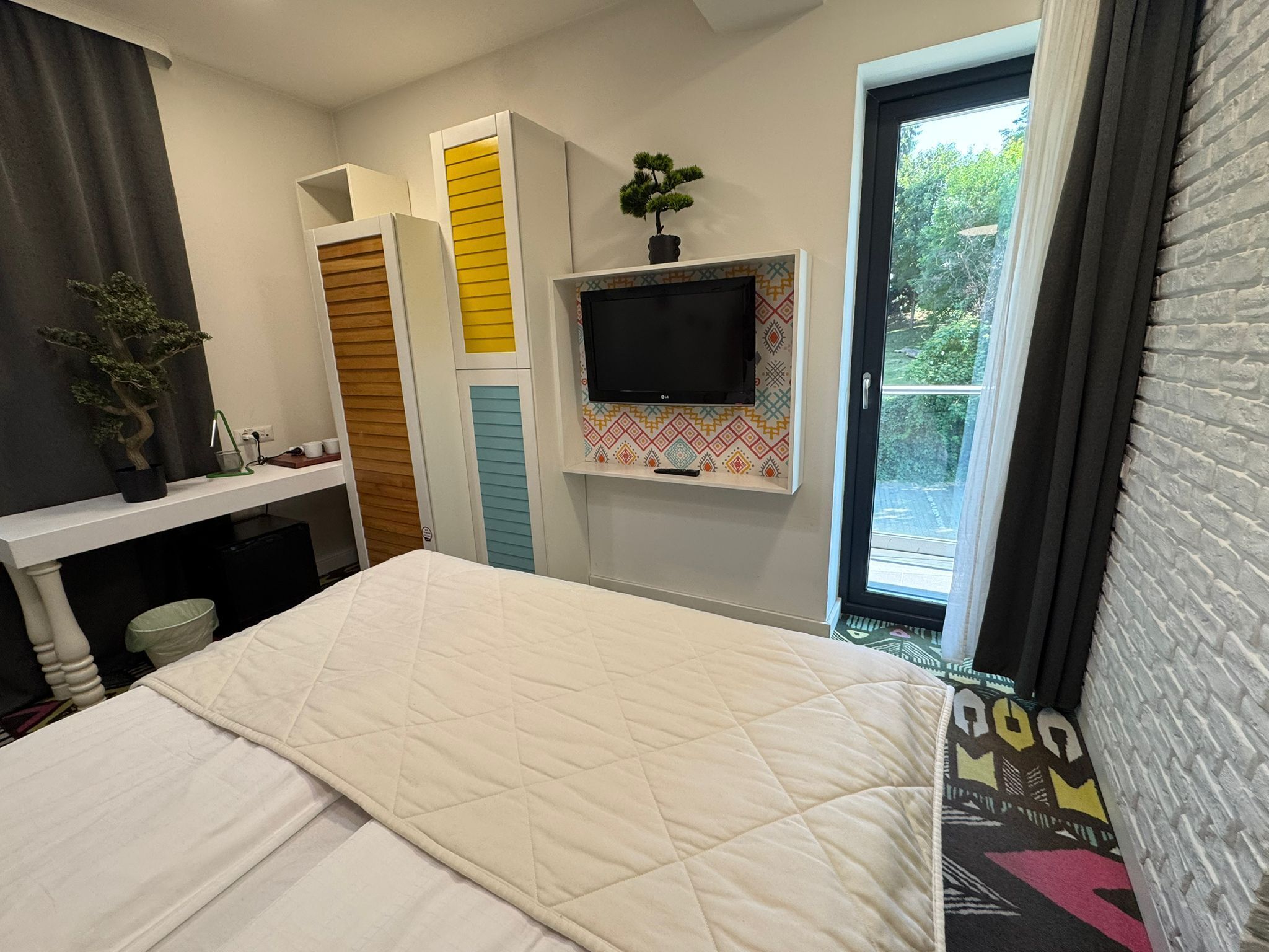 A bedroom with a bed, a TV mounted on the wall, a window with curtains, and a small potted plant on the dresser.