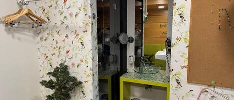 A bathroom with floral wallpaper, a potted plant, a yellow countertop, a mirror, and a corkboard on the wall.