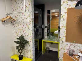Un baño con papel tapiz floral, una planta en maceta, encimera amarilla, espejo y un tablón en la pared.