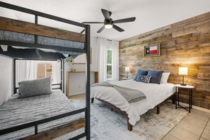 A bedroom with a bunk bed, a single bed, a ceiling fan, a wooden wall, and a flag decoration. - Canyon Cottage: Hill Country Getaway, Hot Tub & Fire Pit, Mins to Whitewater Amphitheater & Tubing (Canyon Lake)