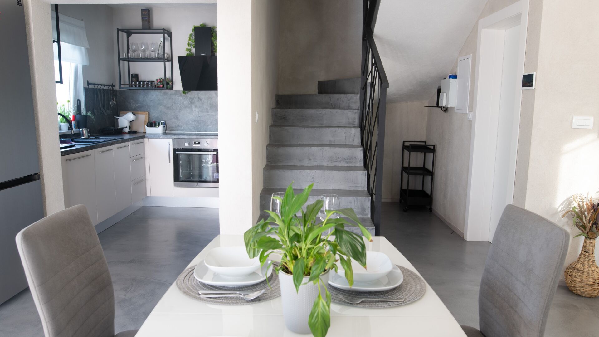 Una cocina moderna con horno empotrado, una zona de comedor con una mesa dispuesta para dos personas, y una escalera que sube a otro nivel.