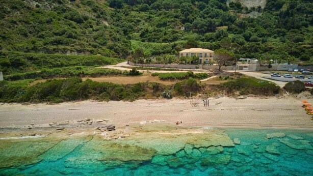 Luchtbeeld van een kustbezit met een groot huis, omgeven door begroeiing, een zandstrand en helder turquoise water.