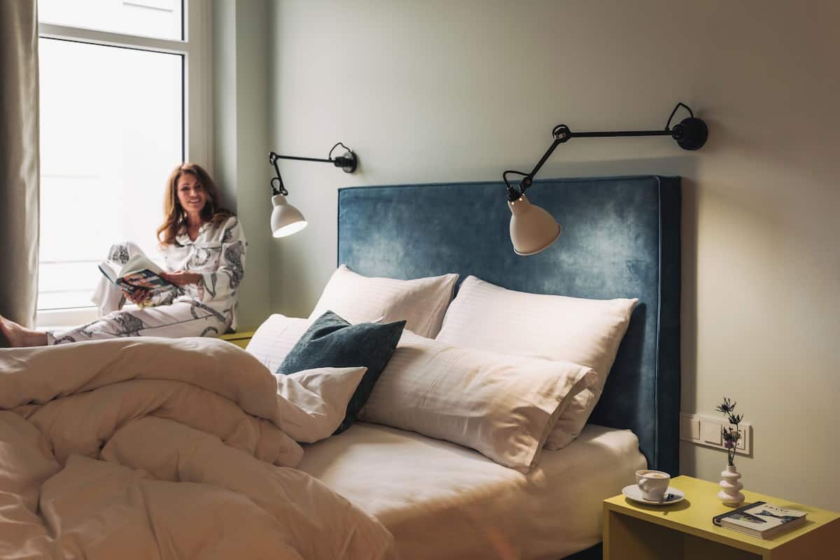 Une chambre à coucher avec un lit, des oreillers, une table de chevet sur laquelle se trouvent une tasse et un livre, et une fenêtre laissant entrer la lumière naturelle.
