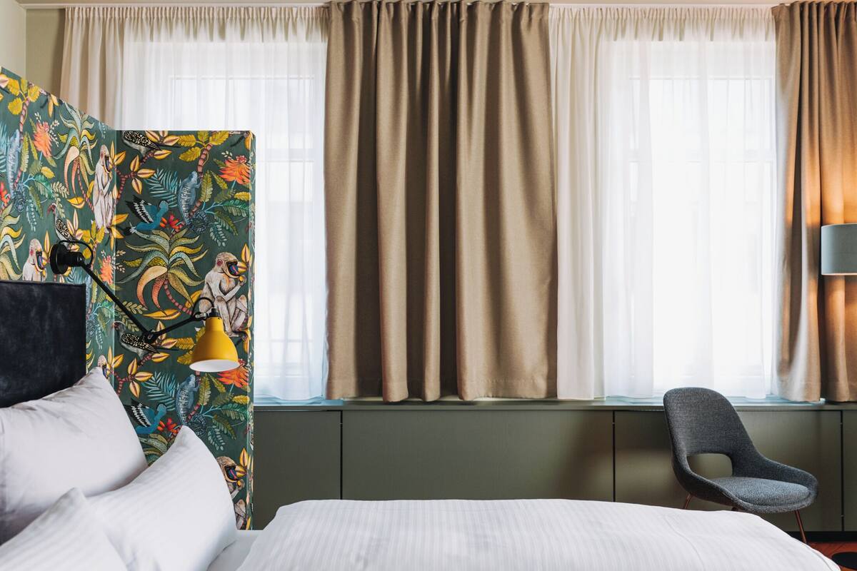 Une chambre d’hôtel avec un lit, une chaise, un bureau avec une lampe et une fenêtre avec des rideaux.