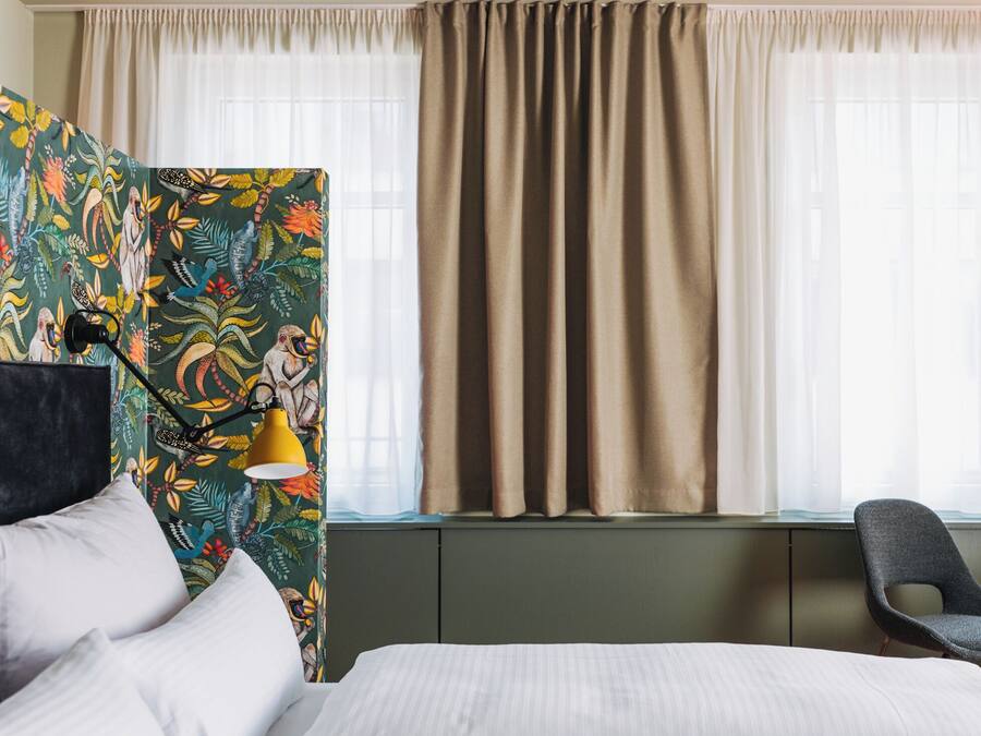 A hotel room with a bed, a chair, a desk with a lamp, and a window with curtains.