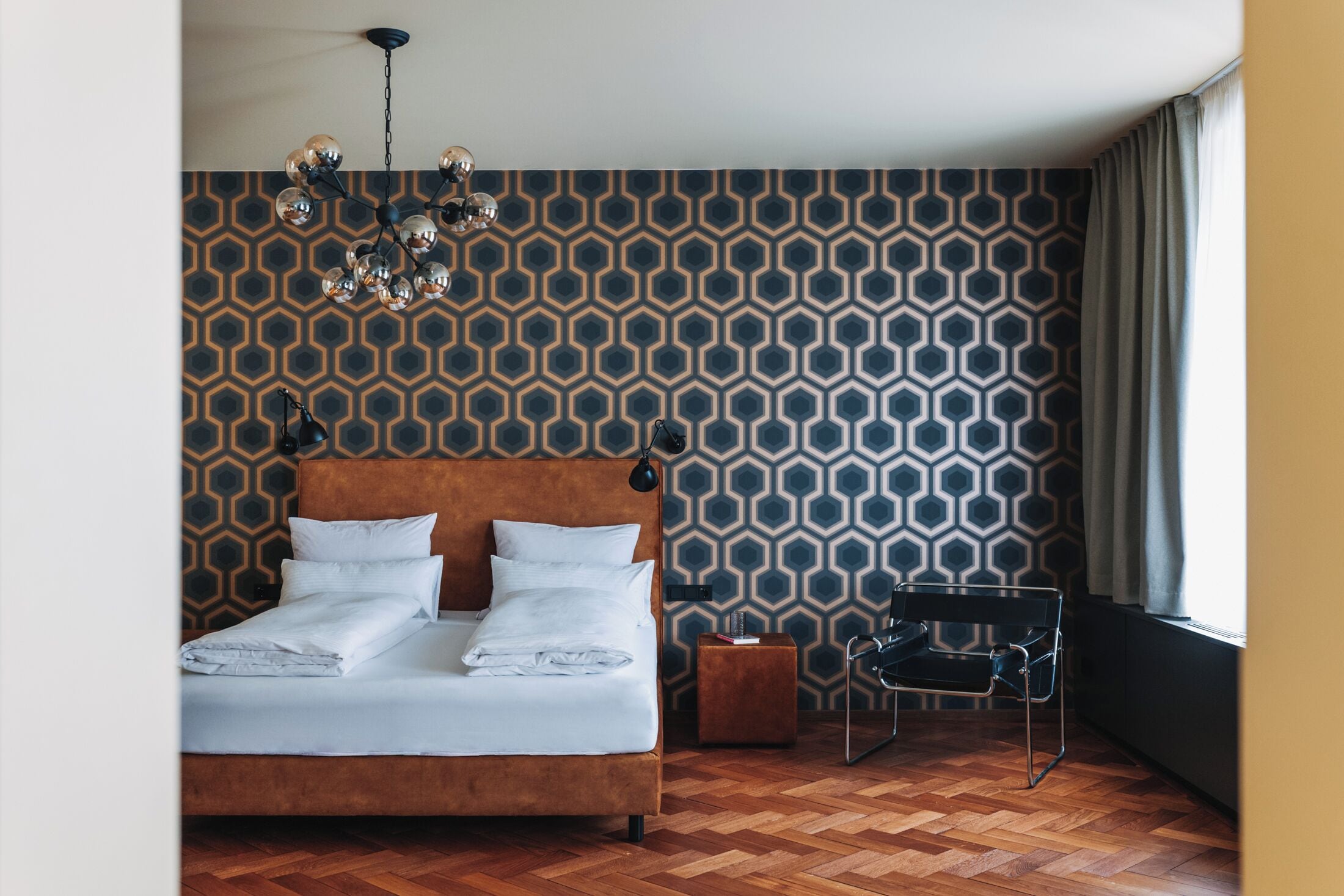 A bedroom with a wooden floor, a bed with white linens, a patterned wall, a hanging light fixture, and a chair by the window.