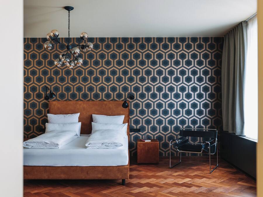 A bedroom with a wooden floor, a bed with white linens, a patterned wall, a hanging light fixture, and a chair by the window.