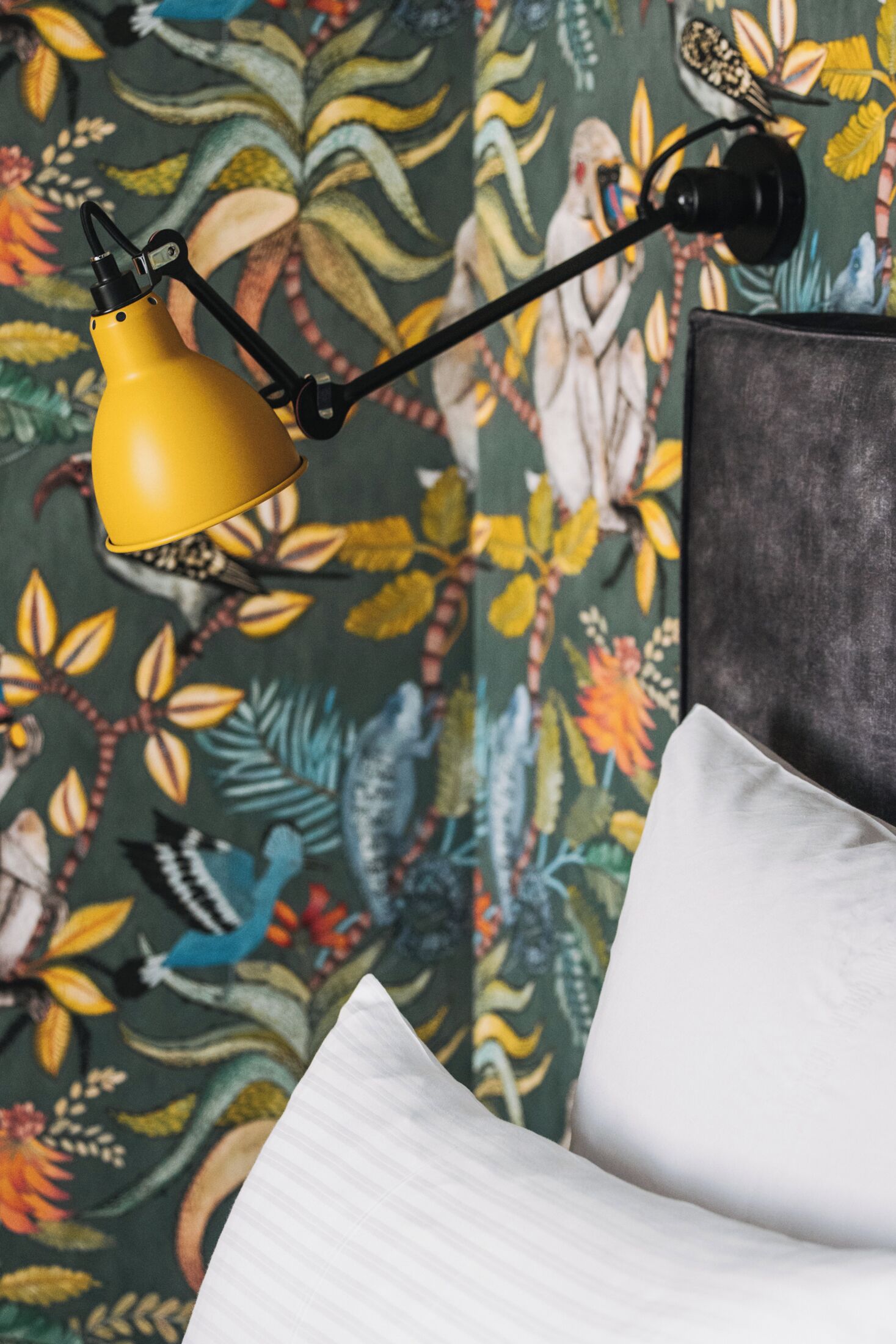 Una cama con un almohadón blanco y una lámpara amarilla sobre un papel tapiz con estampado floral.