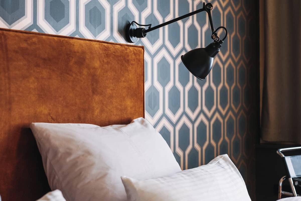 Un lit bien fait, avec une tête de lit en bois, des oreillers blancs et un mur au fond orné d’un motif géométrique.