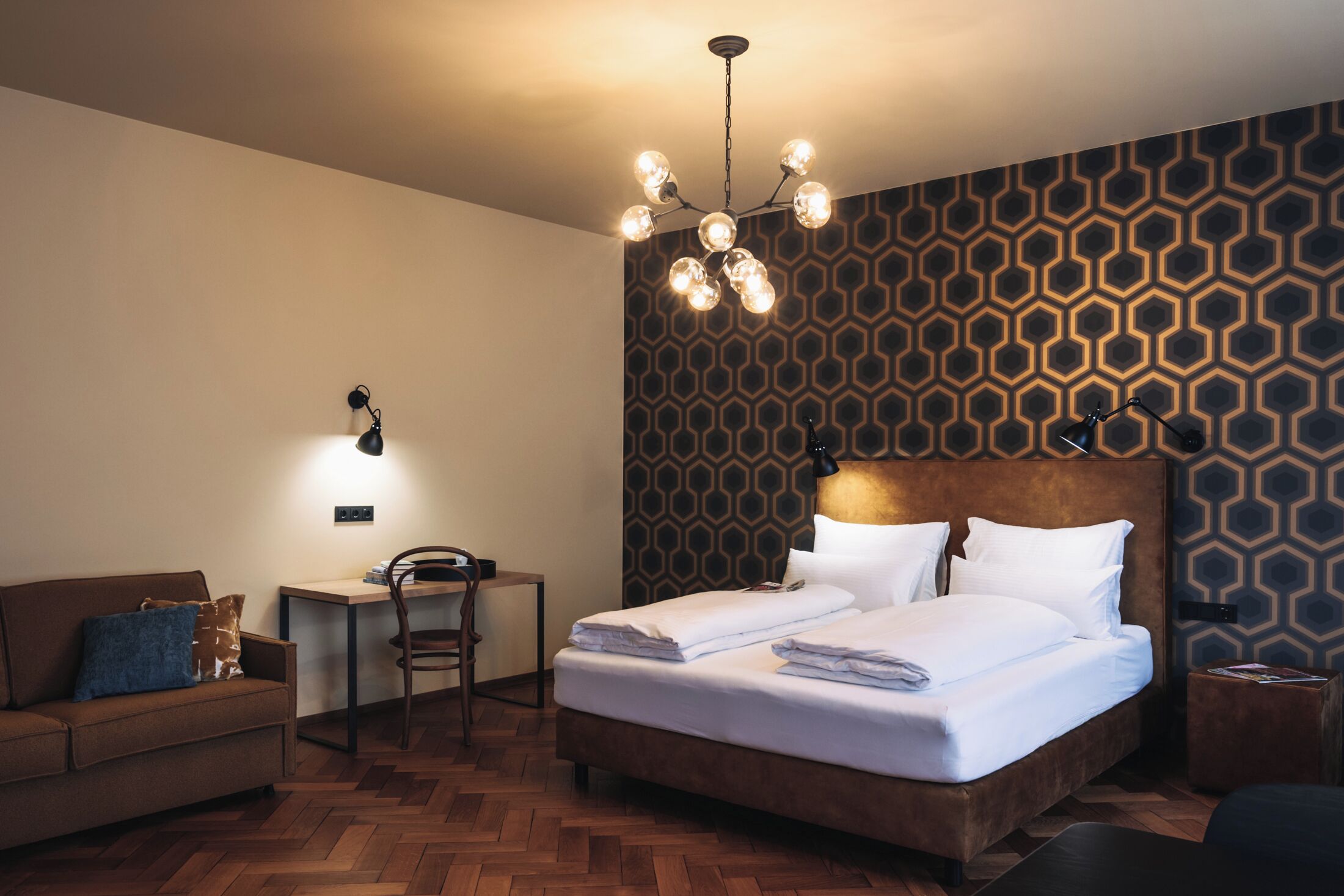 Habitación de hotel con suelo de madera tipo espiga, cama con ropa blanca, cabecero marrón, escritorio con silla y pared con estampado.