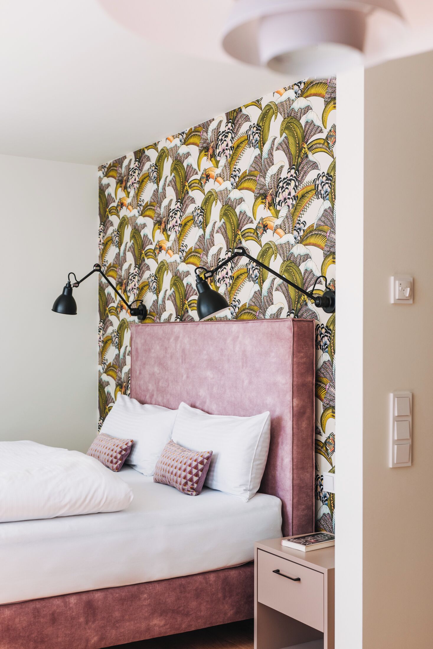 A bedroom with a pink headboard, white bedding, and a patterned wallpaper. There are two black desk lamps on the wall and a bedside table with a book.