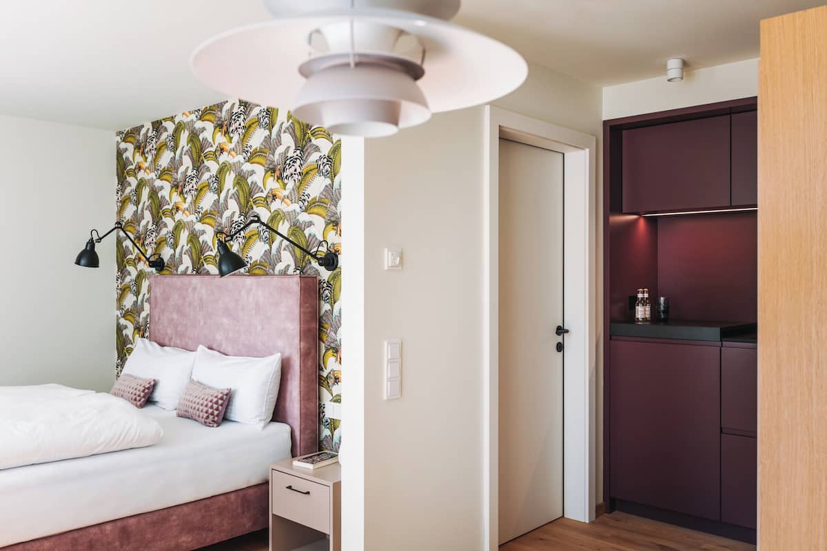 Une chambre à coucher comprenant un lit, une table de chevet et une commode. Une porte donne accès à une cuisine équipée d’armoires et d’un plan de travail.