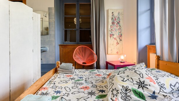 Une chambre à coucher comprenant un lit, une chaise, une petite table avec une lampe, une fenêtre avec des rideaux et une œuvre d’art accrochée au mur.