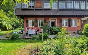 Ein zweistöckiges Gebäude mit einem Garten und einer Terrassenförmig angelegten Sitzgelegenheit, an der Tische stehen.