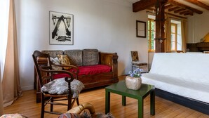 Ein gemütliches Zimmer mit Holzboden, einem grünen Tisch, einem weißen Bett, einer Holzsofa mit Leopardenmuster-Kissen, einem Holzstuhl und einem Bild an der Wand.