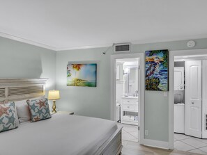 Une chambre à coucher comprenant un lit, une table de chevet, une lampe, un tableau, une salle de bain avec un miroir et une porte donnant sur une autre pièce.