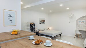 A modern, well-lit room with a wooden dining table set with croissants and coffee cups, a bed with a white mattress and pillows, a small nightstand with a lamp, and a world map on the wall. - Sunny Palace Studio 3 (Dubrovnik)