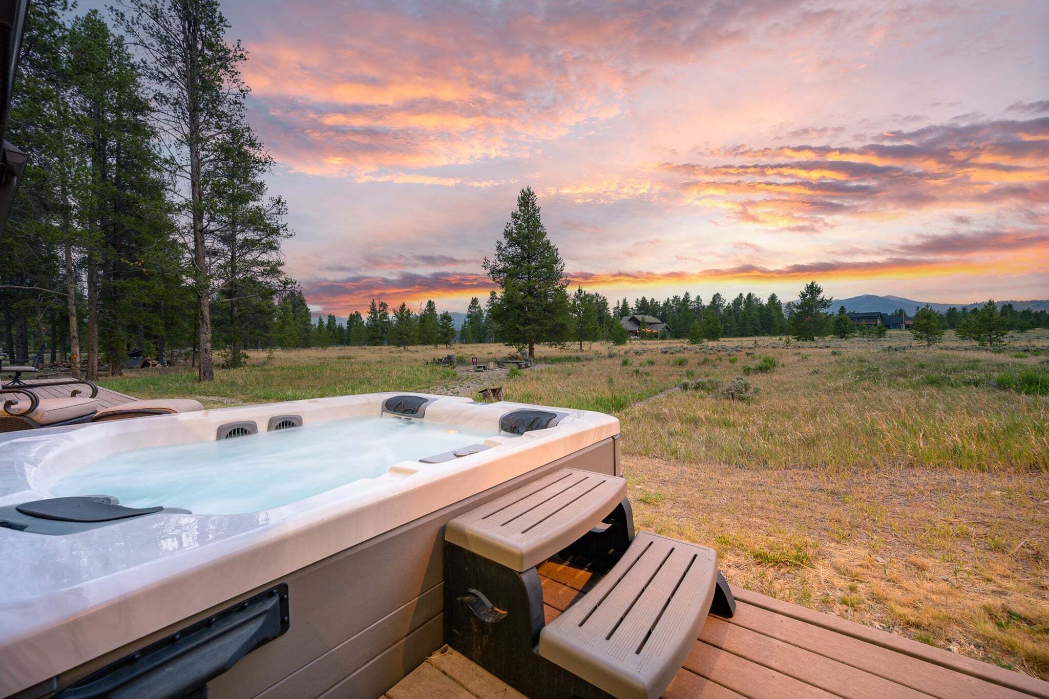 Un bain à remous doté d’une terrasse en bois, entouré d’un champ verdoyant et d’arbres, avec un coucher de soleil en arrière-plan.
