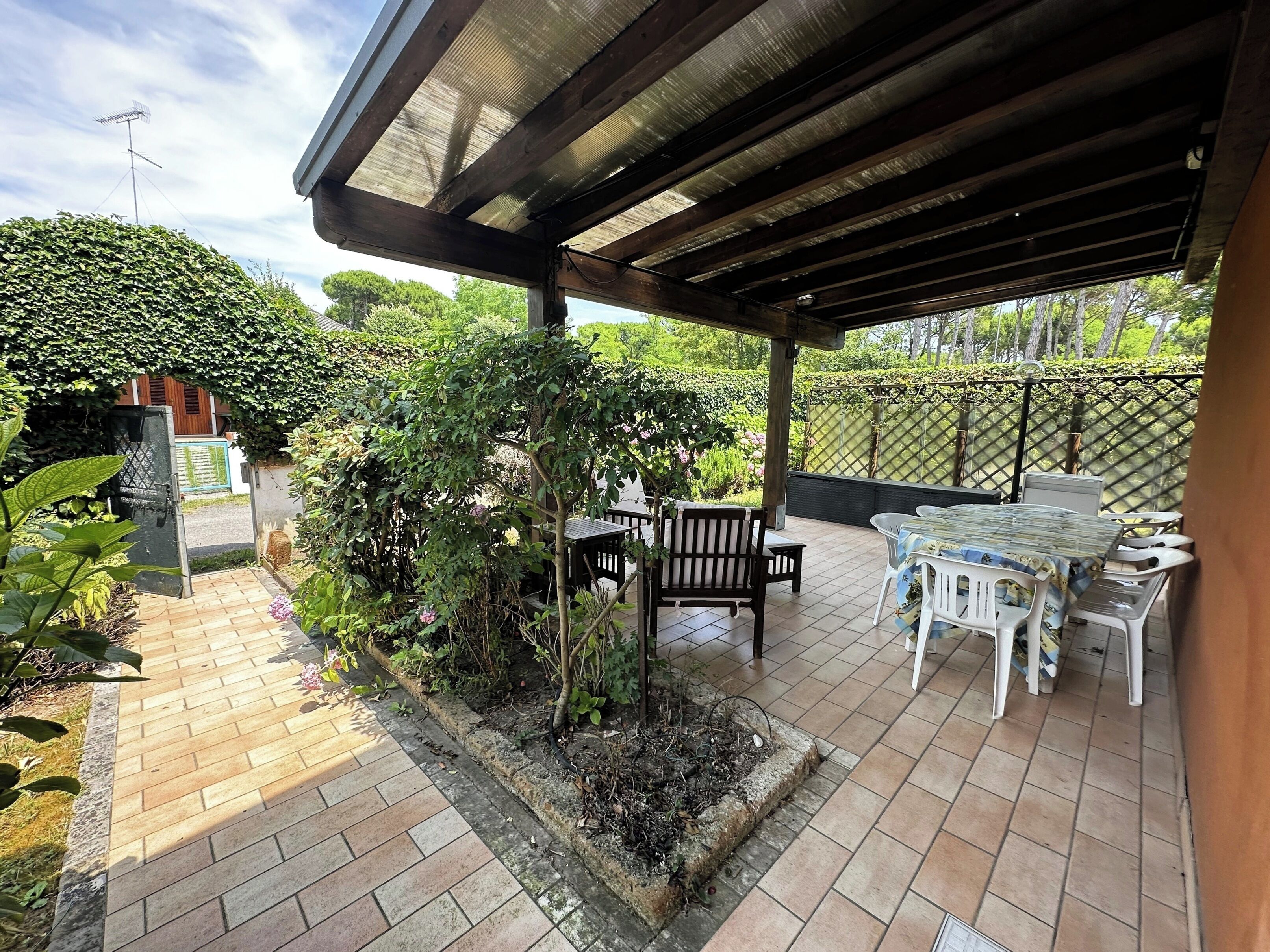 Un patio cubierto con suelo de ladrillo, mobiliario de madera y un área ajardinada con plantas y una cerca.