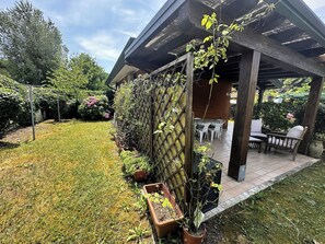 A covered patio area with a table and chairs, surrounded by a garden with various plants and a wooden fence. - Quiet Gardened Villa for six Guests Near the sea (San Michele al Tagliamento)