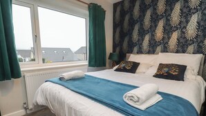 Ein ordentlich bezogenes Bett mit blauer Decke und weißen Kissen, ein Fenster mit grünen Vorhängen und eine Wand mit Muster-Tapete.