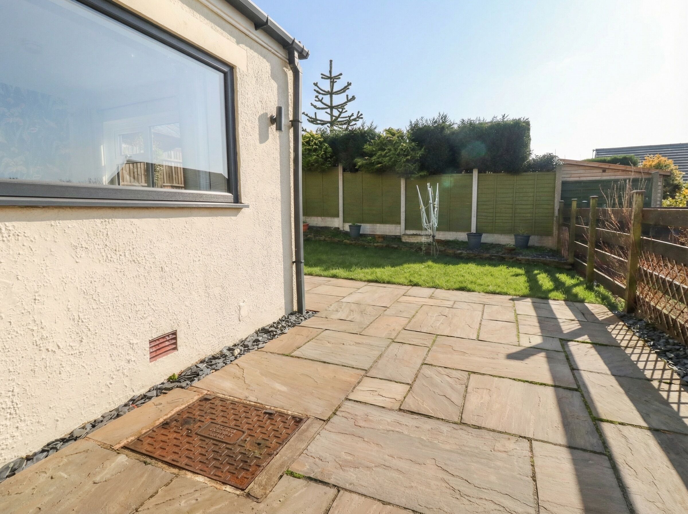 Een woonhuis met een geplaveid terras, een metalen putdeksel, een houten hek en een groen gazon.